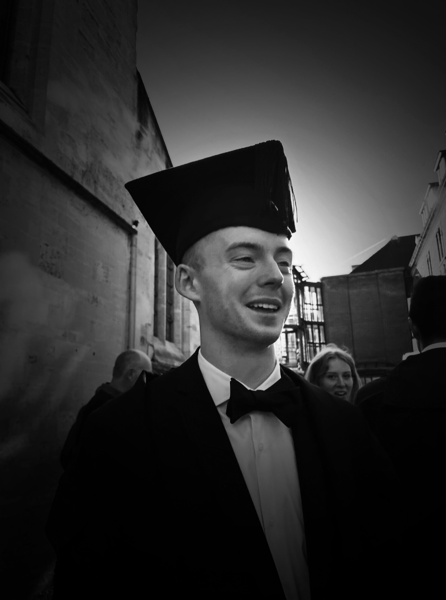 A young man in formal attire with a bow tie and academic cap, smiling at a graduation ceremony outdoors in black and white.