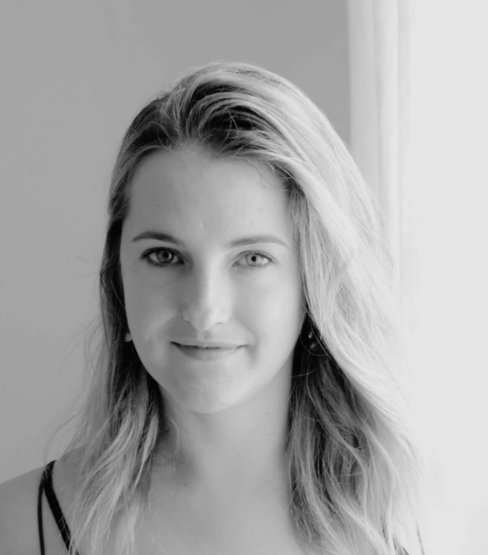 A black-and-white portrait of a young woman with long wavy hair, smiling gently, standing near a window.