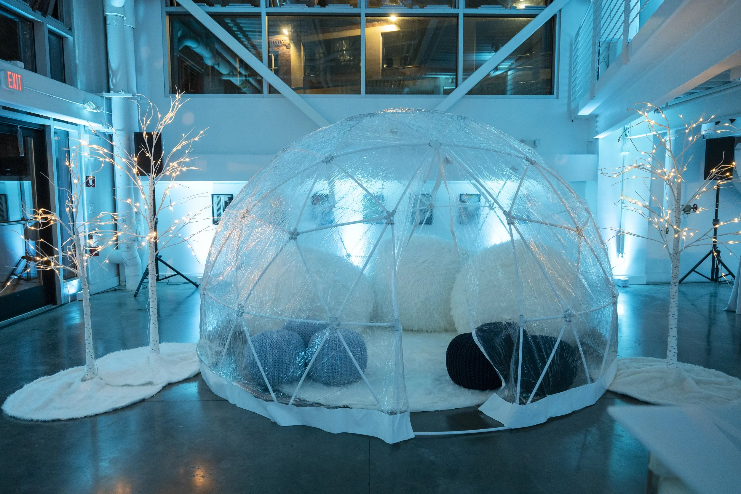Clear igloo lounge with white and knit poufs between glowing birch trees under ice-blue light at a Boston winter holiday party at Artists For Humanity, produced by Siagel Productions