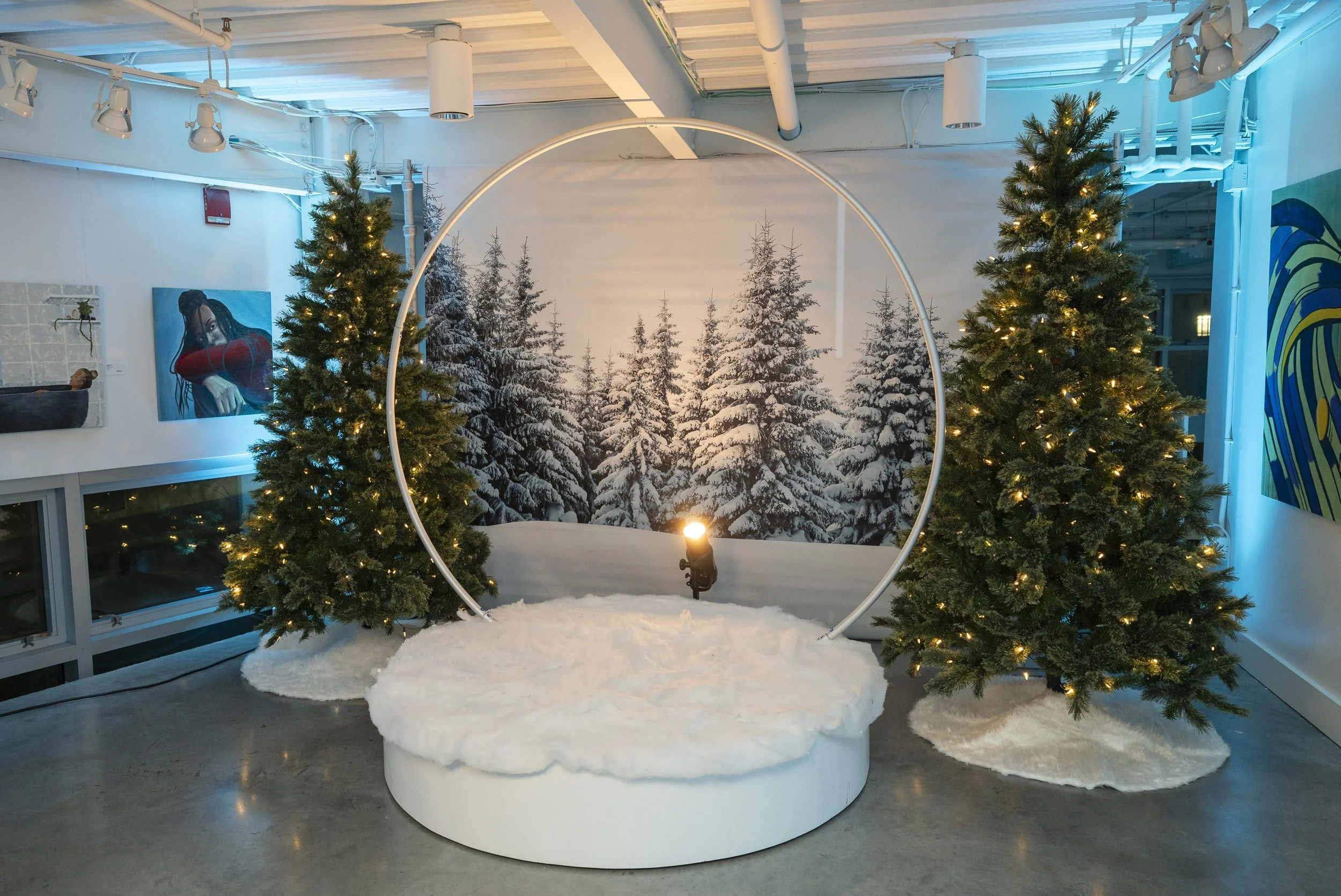 Winter-themed photo booth with a snowy pine forest backdrop, white circular arch, lit evergreens, and a plush white seat at a Boston holiday party by Siagel Productions