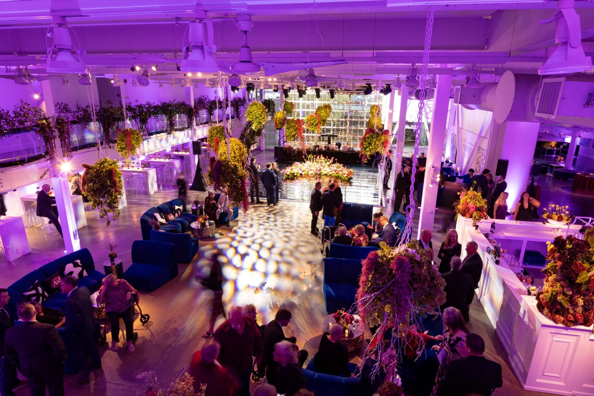 Wedding guests in blue velvet lounges under suspended florals and violet uplighting at Artists For Humanity in Boston, planned and produced by Siagel Productions