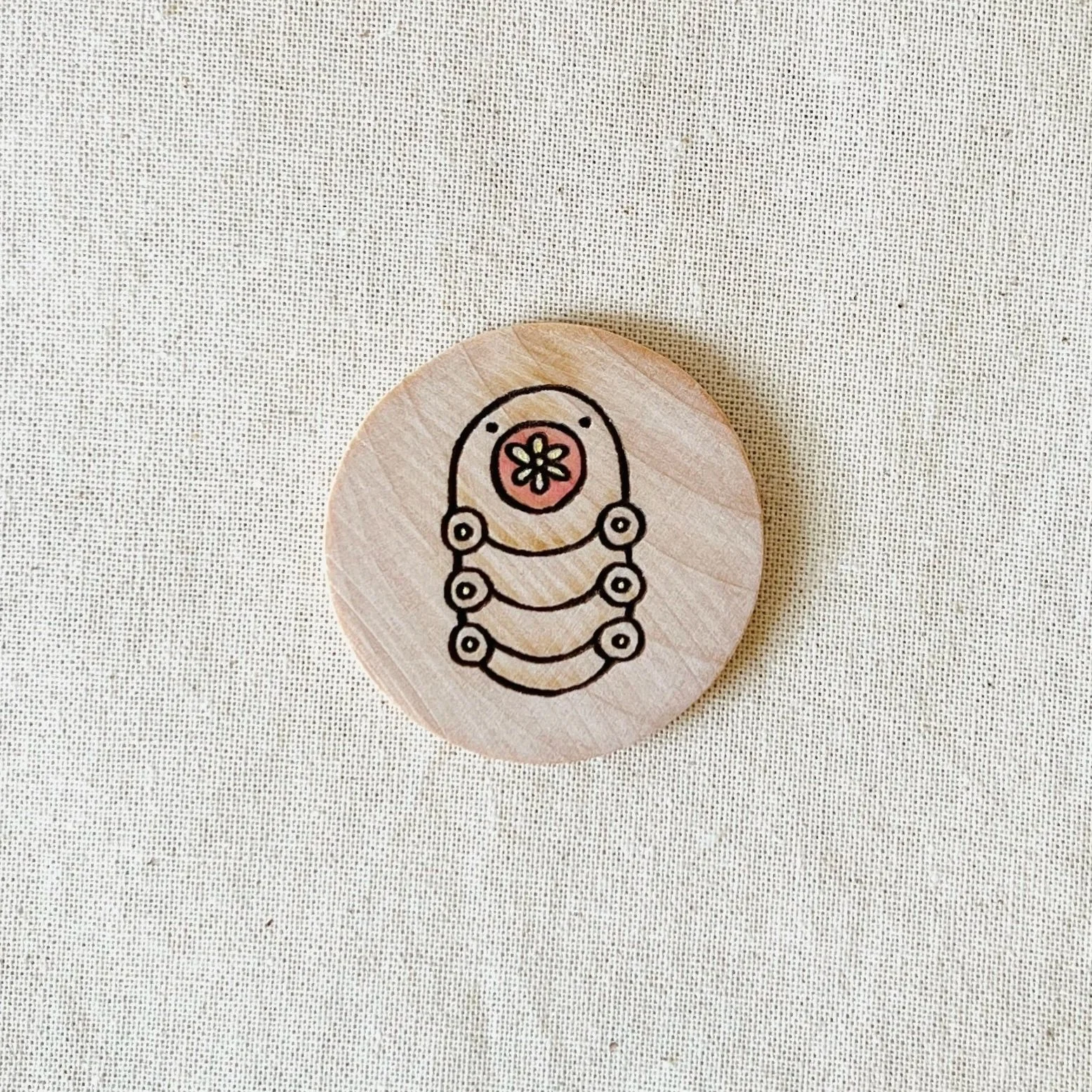 A round wooden compliment coin with a black outline drawing of a stylized insect or caterpillar with a flower on its head, placed on a light-colored fabric surface.