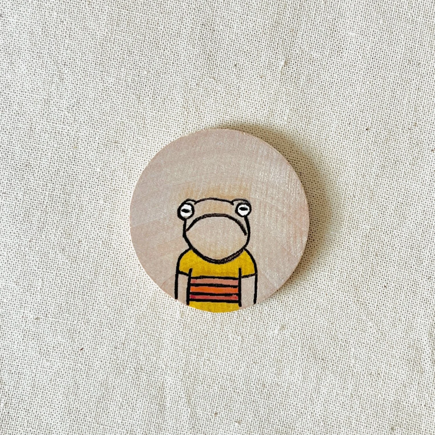 A circular wooden compliment coin featuring a cartoon illustration of an anthropomorphic animal with a large nose, wearing a yellow shirt with red stripes, on a beige background.