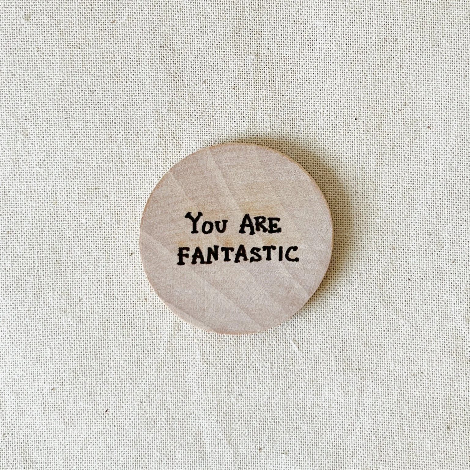 Round wooden compliment coin with the message "You Are Fabulous" in black text on a light-colored background.