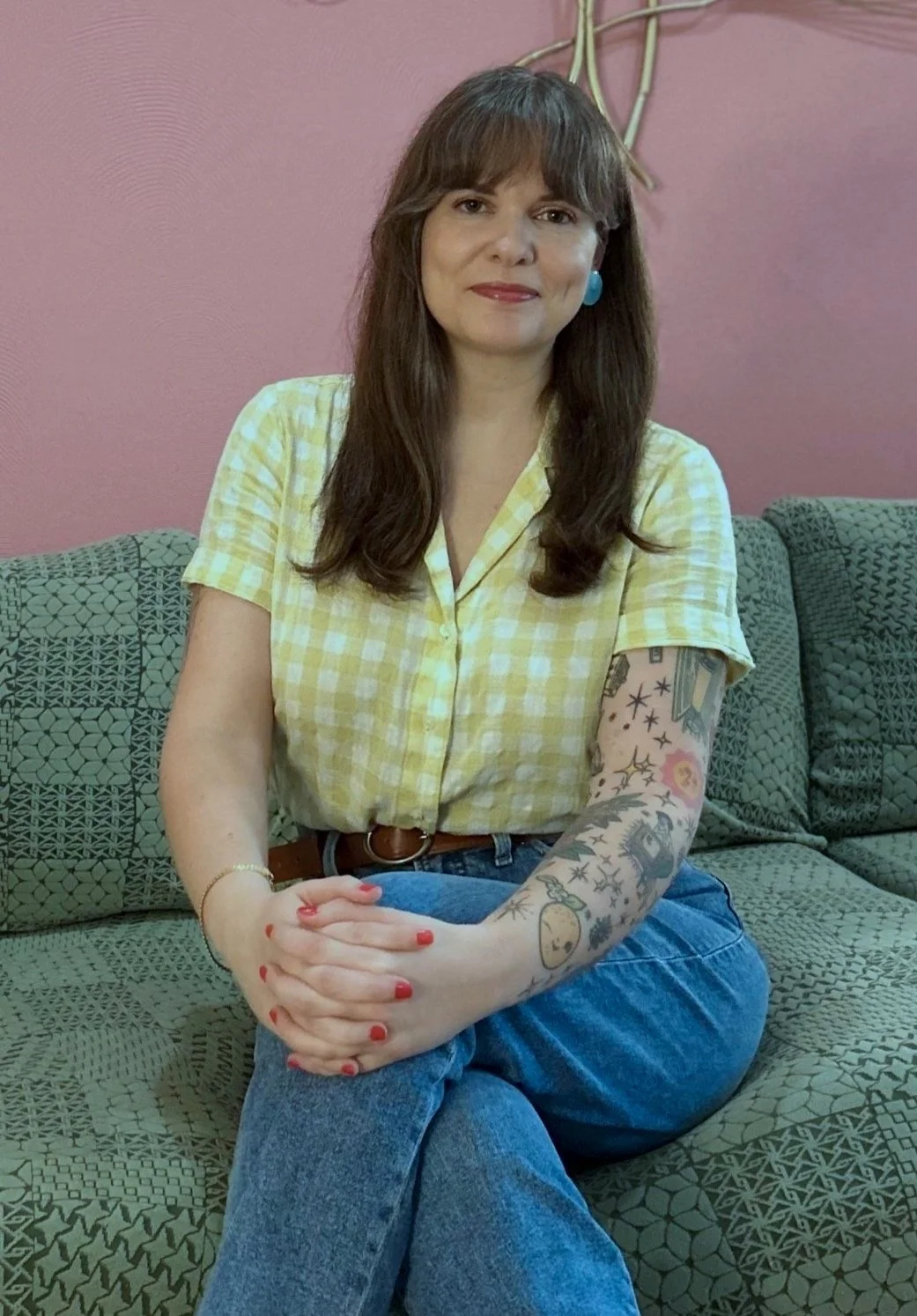 A woman with brown hair and tattoos on her arm, wearing a yellow checkered shirt and blue jeans, sitting on a patterned green sofa against a pink wall.