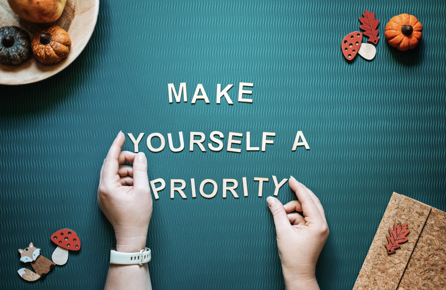 Hands arranging letter tiles on a teal textured surface with the phrase "Make Yourself a Priority." Decorations include small pumpkins, a woodland creature figure, and autumn leaf and mushroom cutouts.