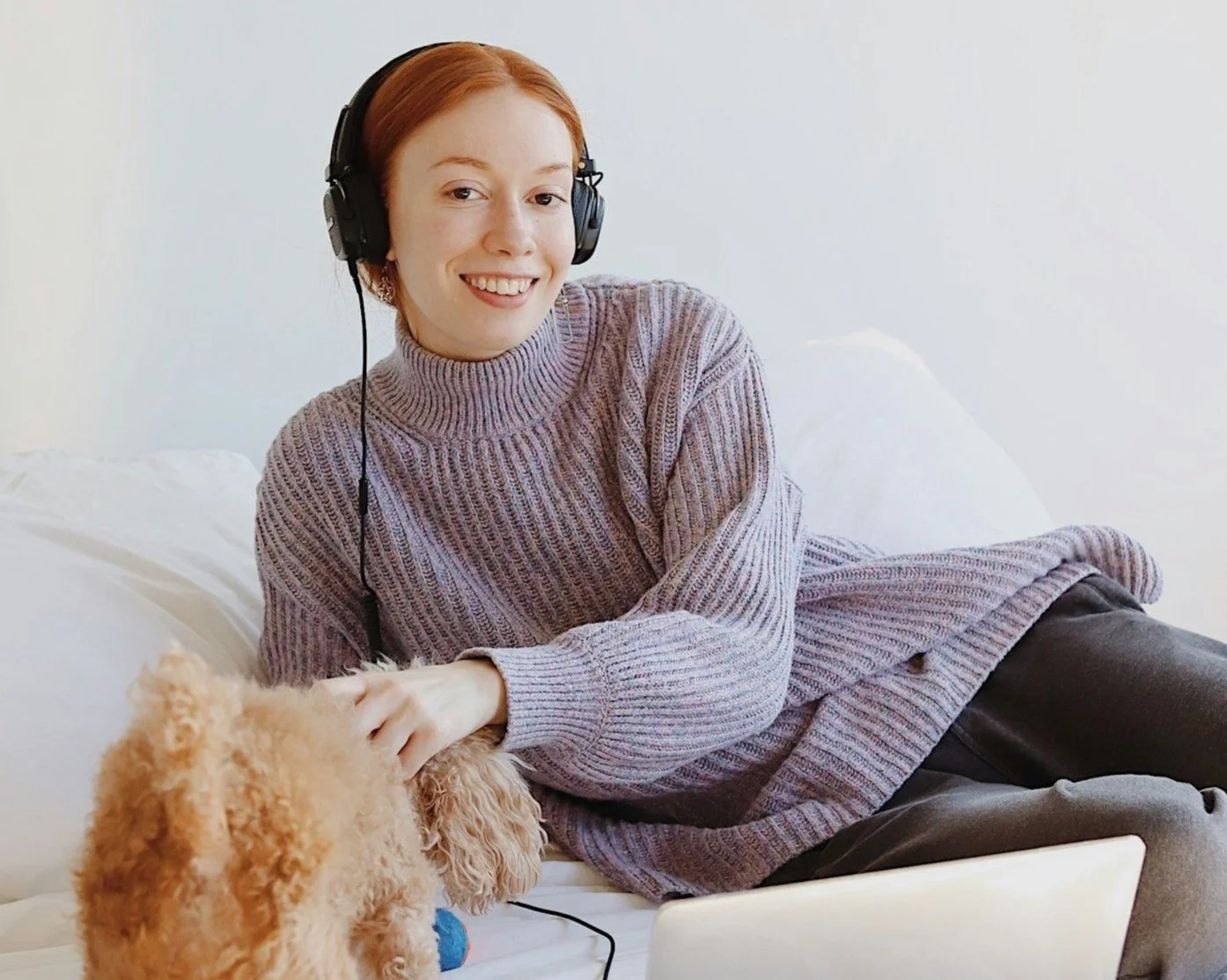 A woman with red hair wearing a purple sweater and headphones lying on a white couch and petting a small, curly-haired tan dog.