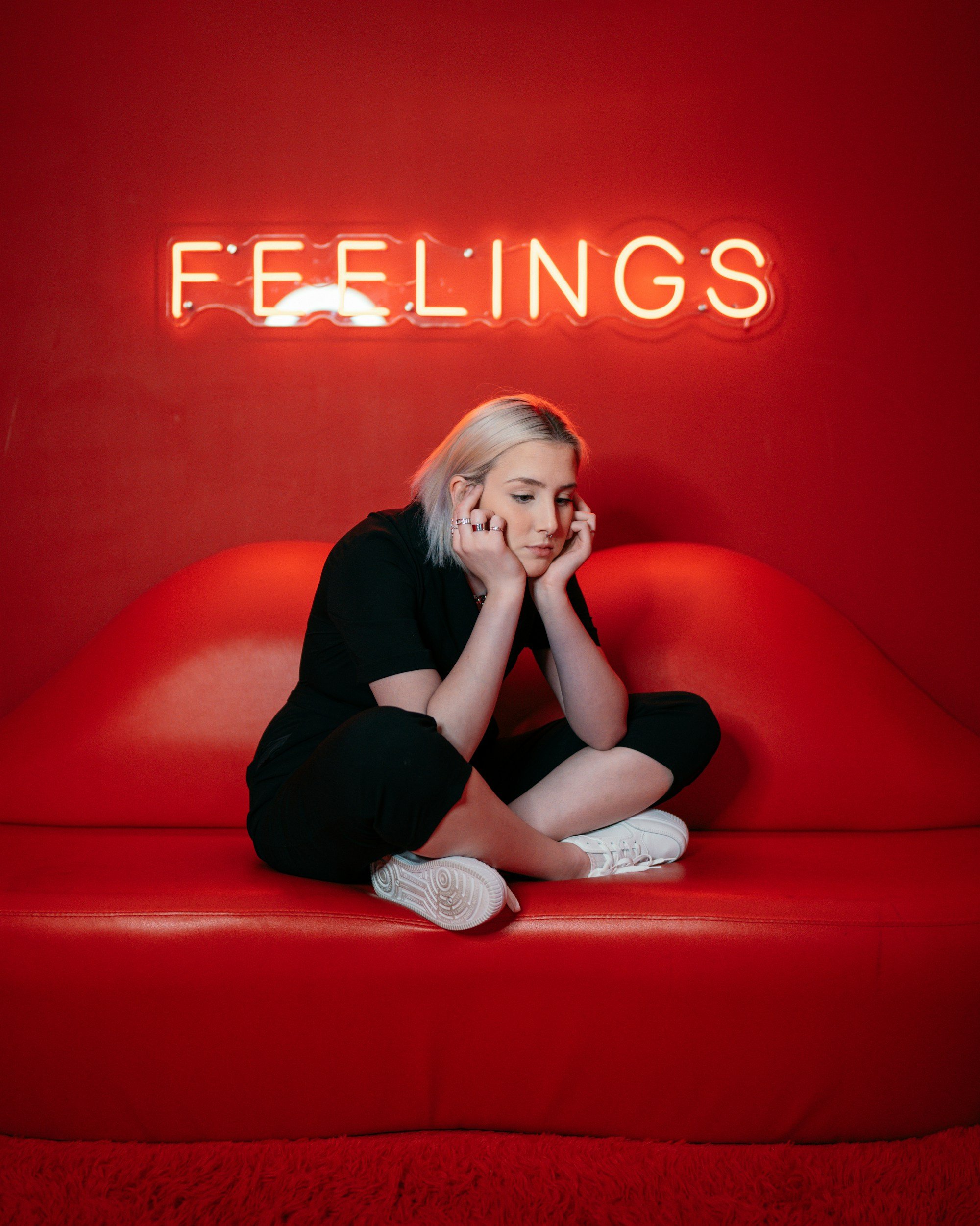 A young woman with platinum blonde hair sitting on a red couch with a neon sign that says 'FEELINGS' behind her. She appears contemplative, resting her chin on her hands.