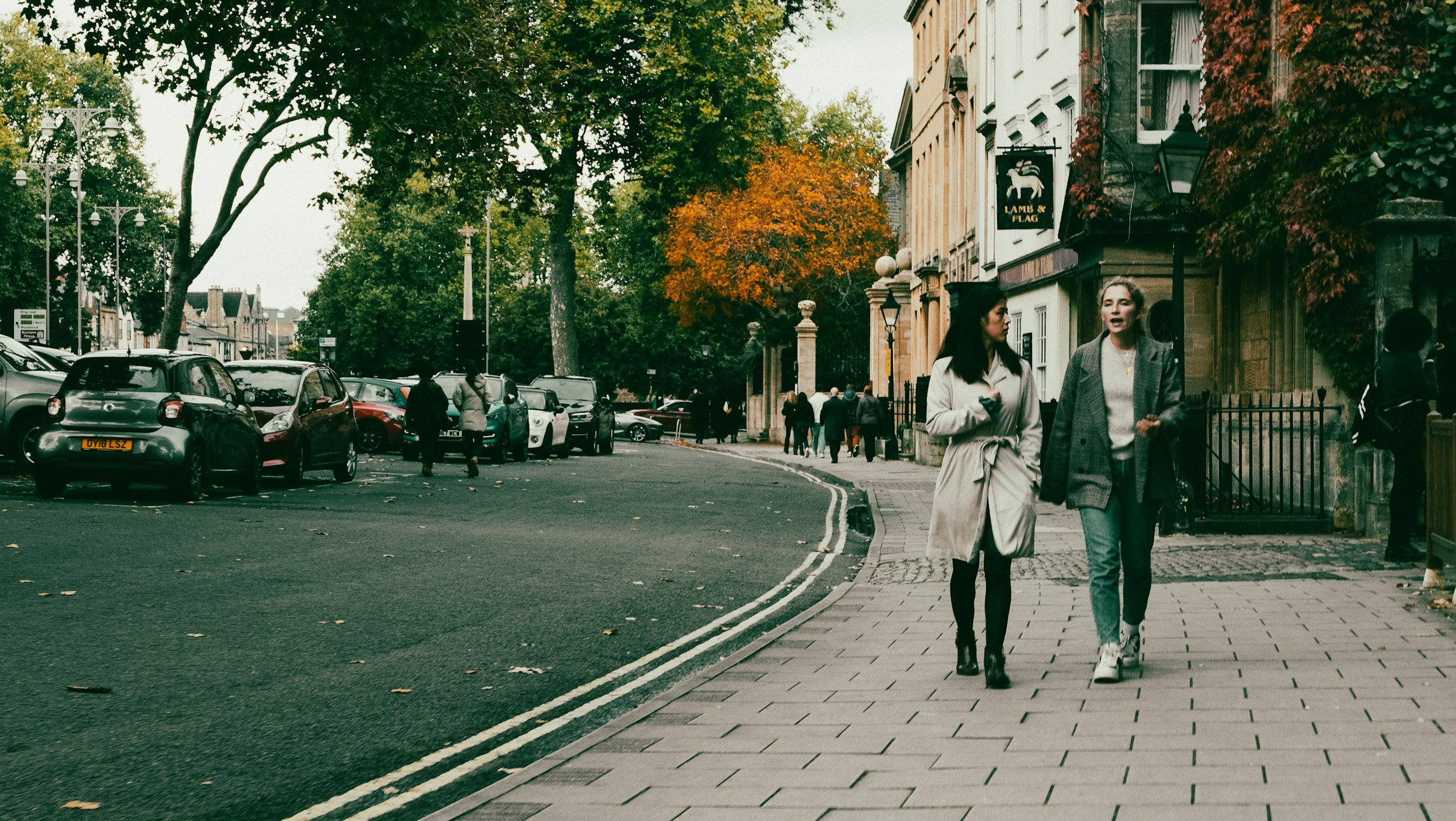 Two women walk and talk along a city sidewalk lined with parked cars and trees with green and orange leaves. In the background, more pedestrians walk past historic buildings with a shop sign for "Lamb & Flag."
