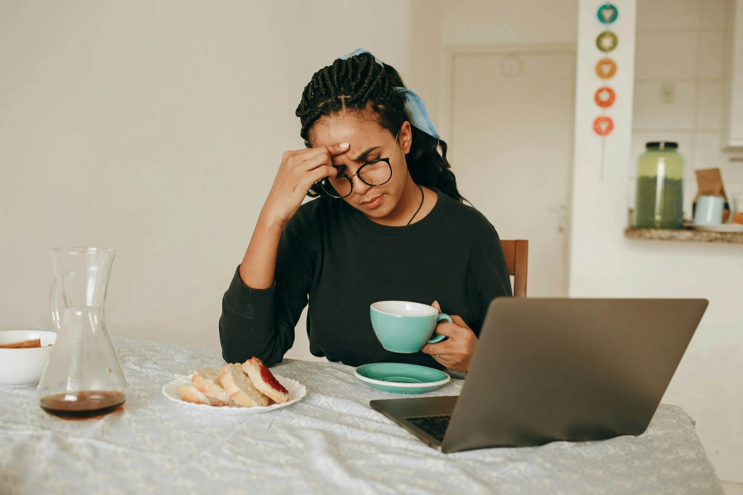 A woman sitting at a table, looking stressed or tired, holding a cup of tea or coffee with a laptop, plate with slices of cake, and a glass pitcher on the table.