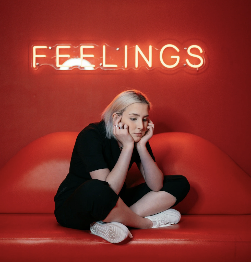 A young woman with short blonde hair, wearing a black outfit and white sneakers, sitting on a red couch with her elbows resting on her knees and her hands supporting her face, gazing downward. Behind her, a neon sign reads 'FEELINGS' on a red wall.