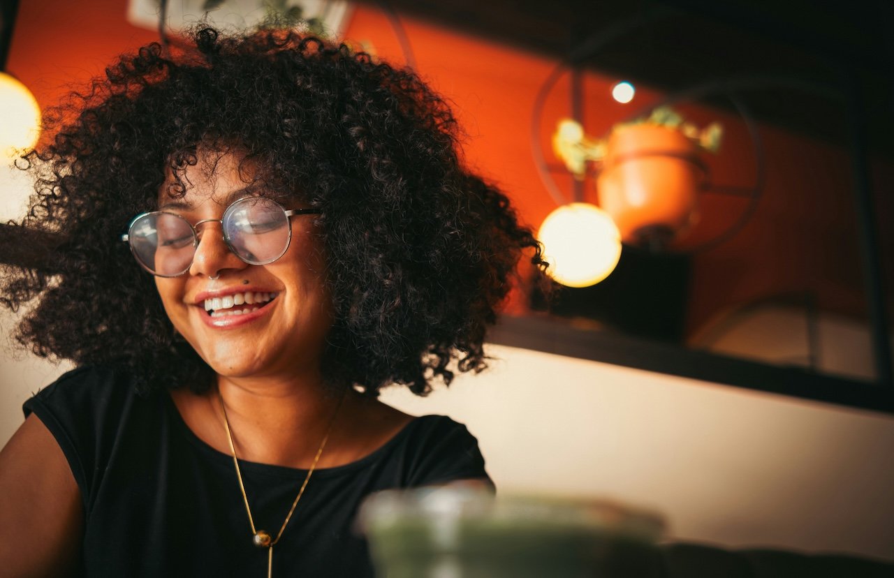A woman with curly hair and glasses smiling in a cozy indoor setting with warm lighting and orange wall decorations.