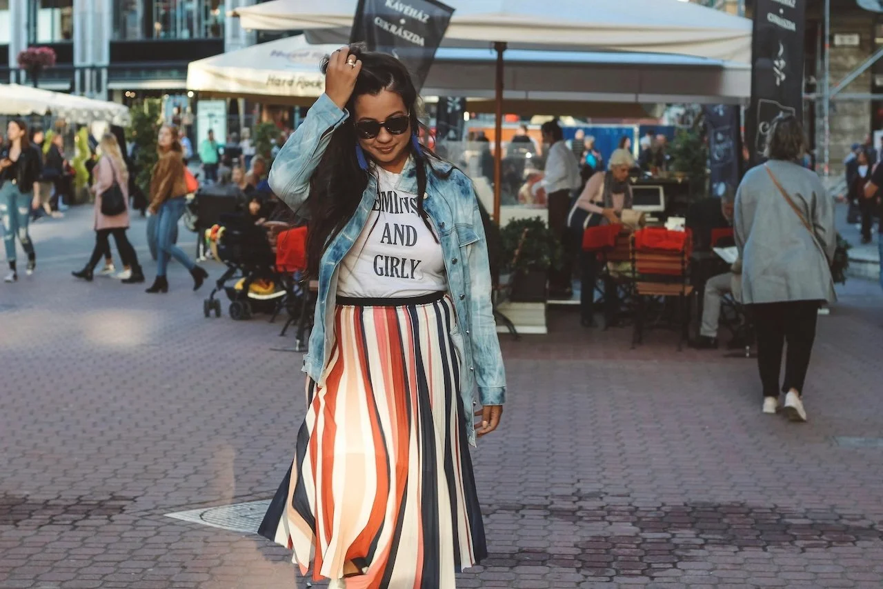 A woman in sunglasses, a denim jacket, a graphic T-shirt, and a long striped skirt standing outdoors on a busy city street, with people and outdoor stalls in the background.
