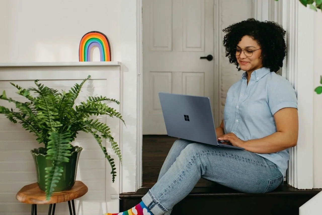 A woman with curly hair and glasses sitting on the floor with a laptop, near a potted fern and a colorful rainbow decoration on a shelf.