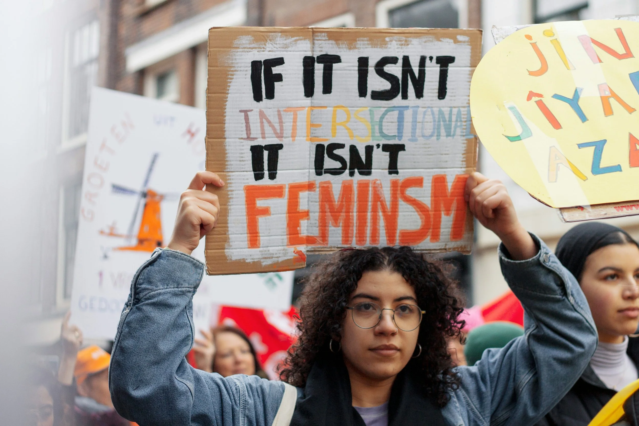 A young woman with glasses and curly hair holding a protest sign that reads, 'If it isn't intersectional it isn't feminism,' during a rally, with other protesters and signs visible in the background.