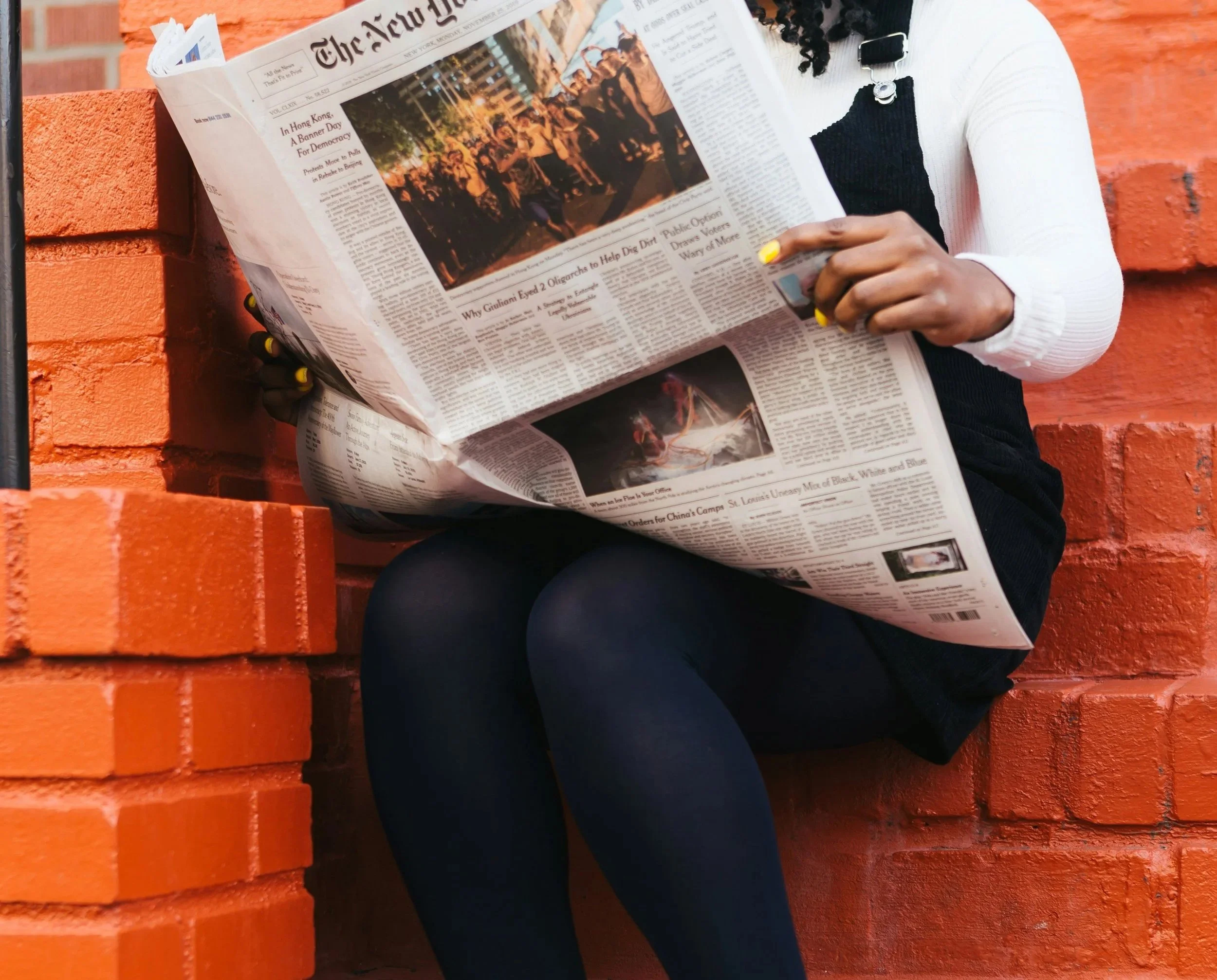 A person sitting against an orange brick wall, reading a newspaper.