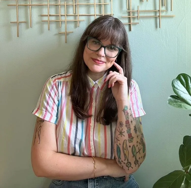 A woman with long brown hair wearing glasses and a colorful striped shirt, standing in front of a light green wall with a wooden wall art piece, and a potted plant on her right side.