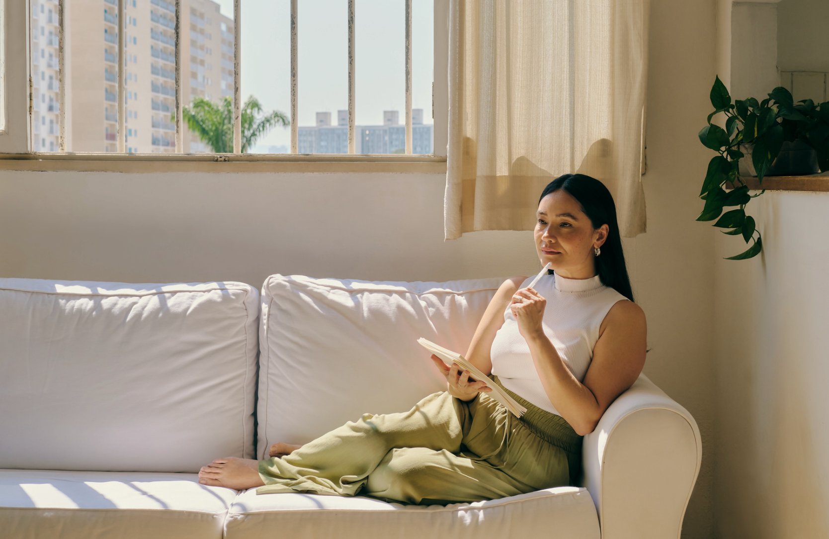A woman sitting on a white couch, holding a notebook and pen, thinking, in a sunlit room with large window and potted plant.