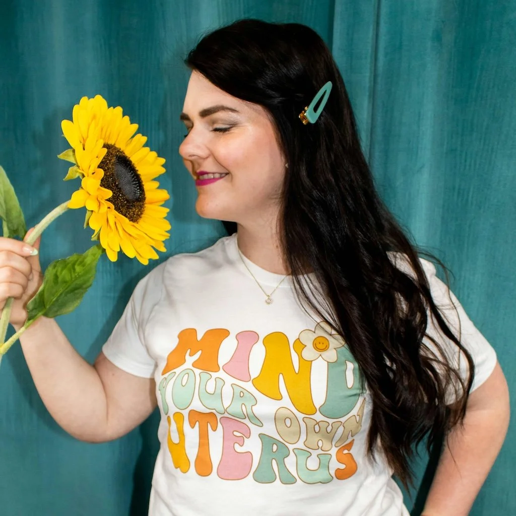 A woman holding a sunflower close to her face, with her eyes closed and a smile, standing against a teal background, wearing a white t-shirt with colorful lettering that says 'MIND YOUR OWN OTERUS' and a small flower graphic.