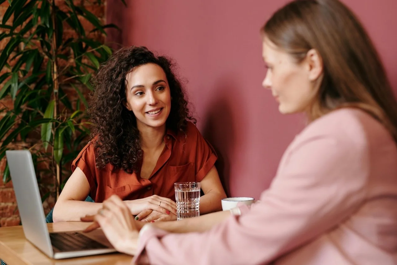 Two women having a conversation at a table, with a laptop, a glass of water, and a mug present, in a cozy indoor setting.