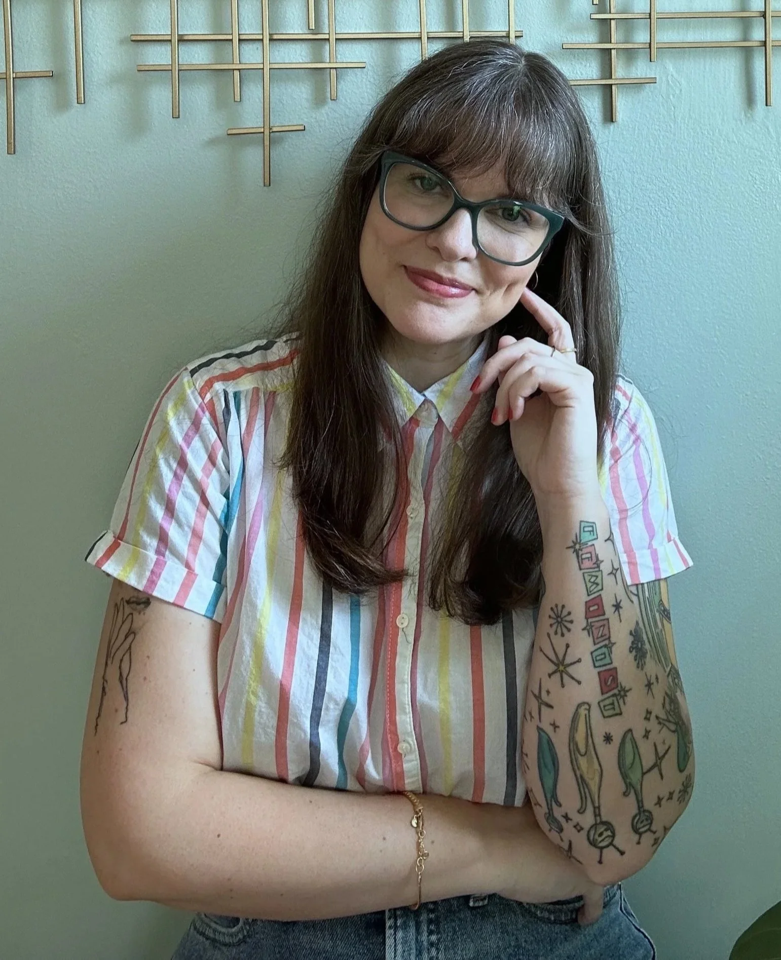 A woman with long brown hair, wearing glasses and a multicolored striped shirt, stands in front of a blue wall with decorative metal rods. She has tattoos on her left arm and is smiling with her hand resting on her cheek.