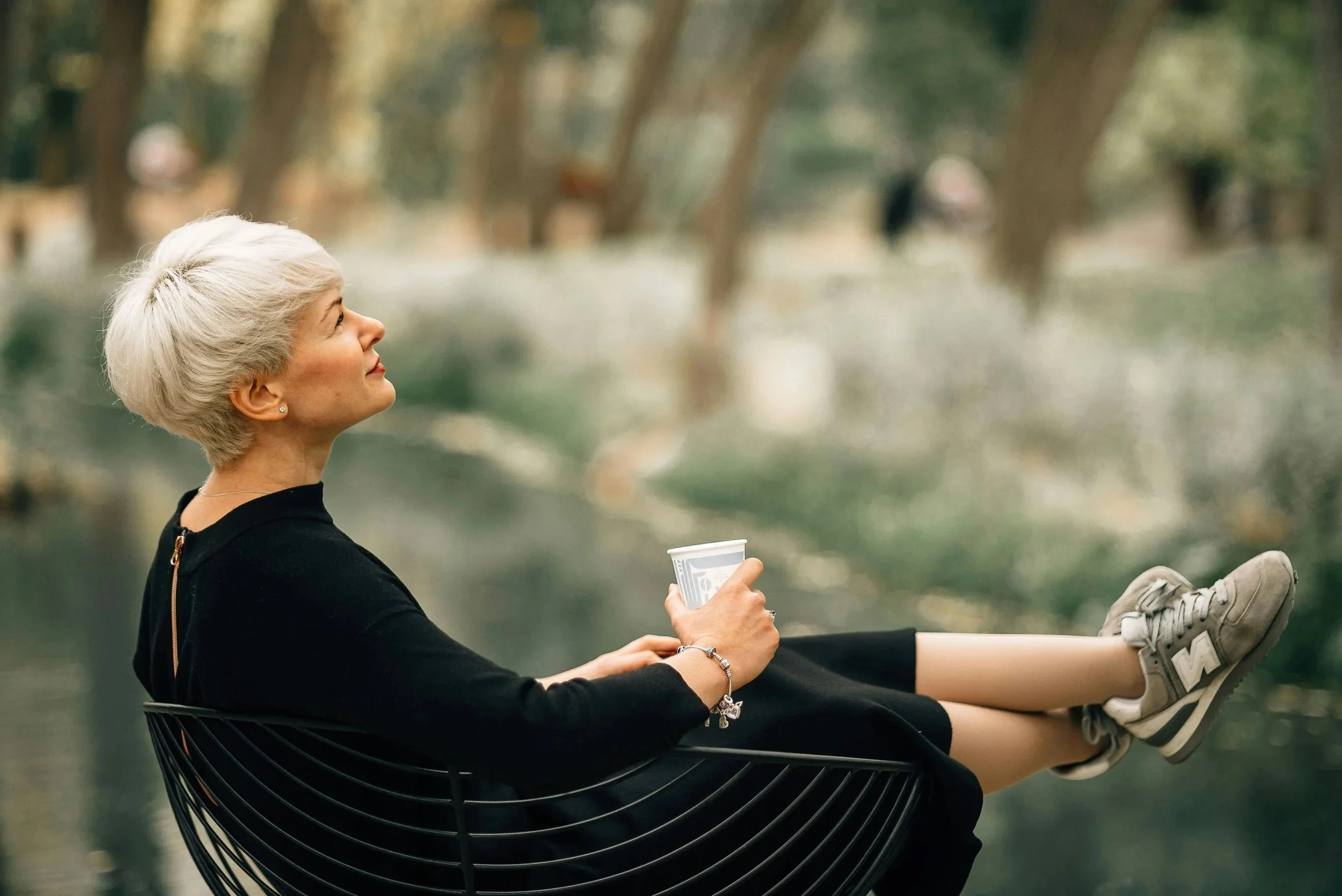 An older woman with short, platinum blonde hair sits in a black outdoor chair by a body of water in a park, holding a paper coffee cup in her right hand and gazing upward with a content expression.