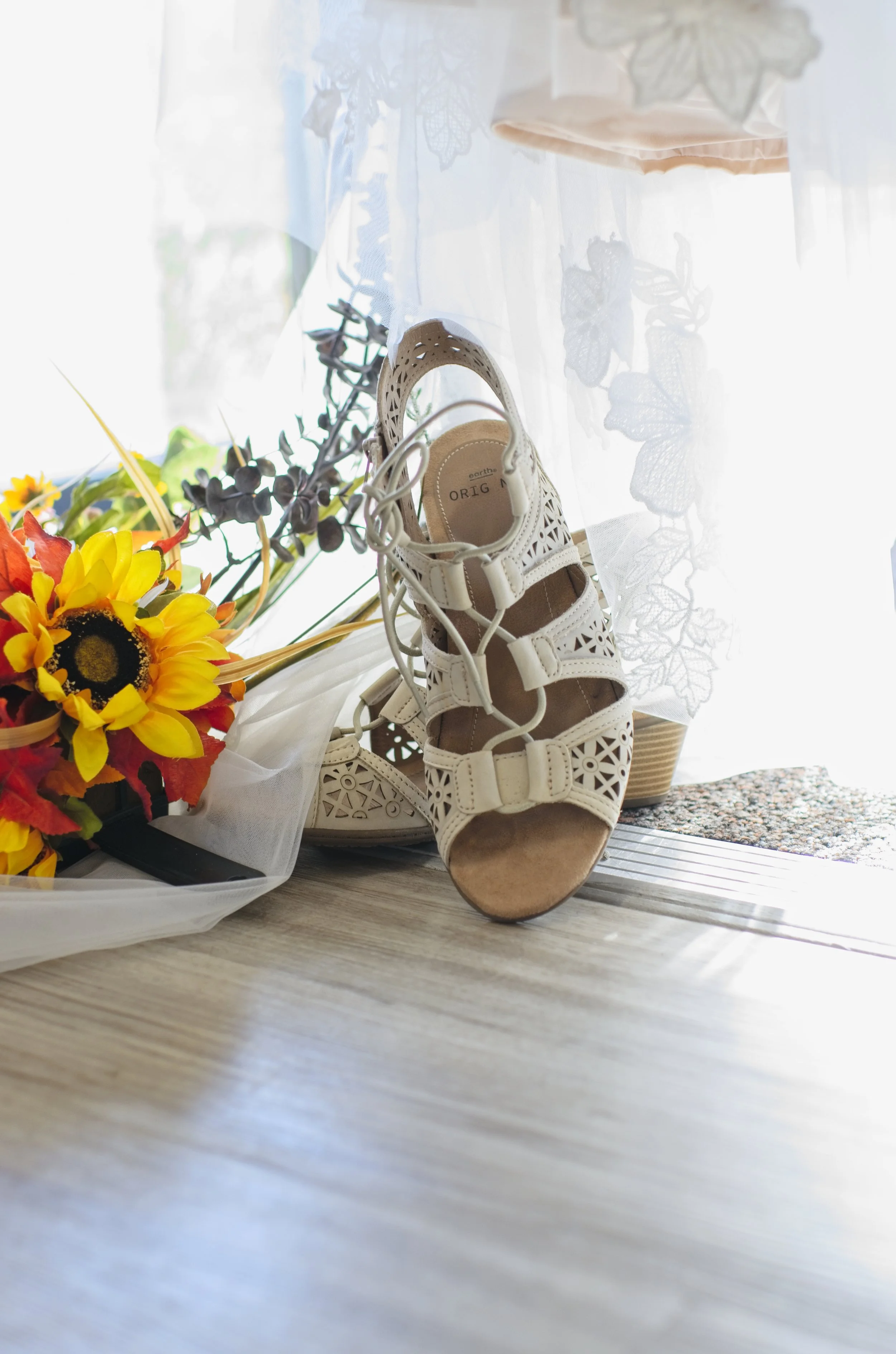 Wedding details include a pair of white lace-up sandals on wooden floor with colorful bouquet and sheer lace curtain.