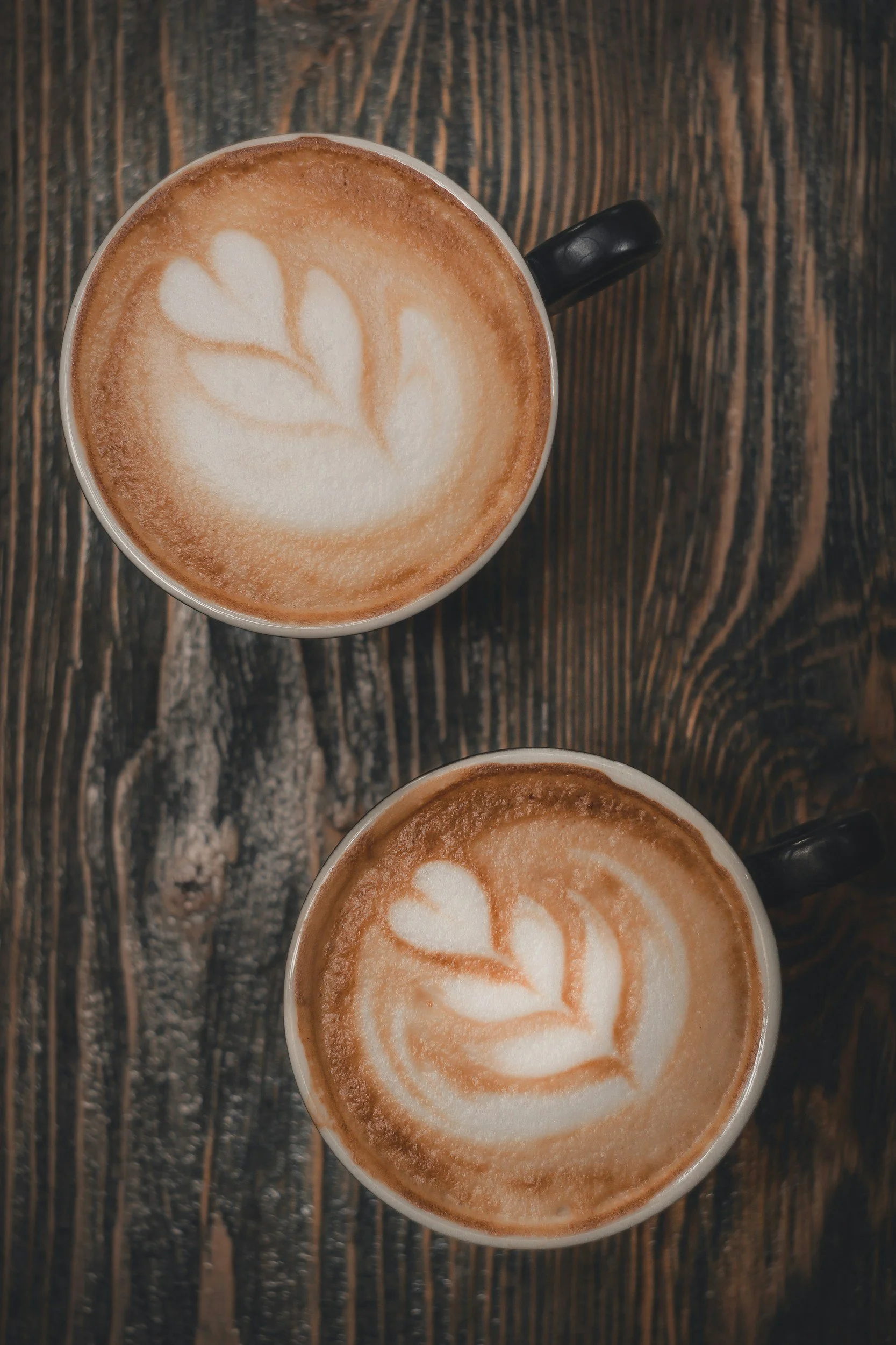 Two cappuccinos sit side by side on a rustic wooden table, showcasing their creamy foam and rich espresso color.