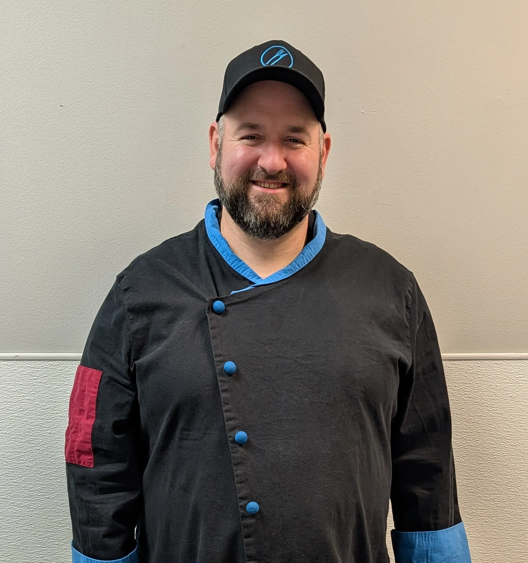 A man with a beard smiling, wearing a black chef jacket with blue accents on the collar, sleeves, and buttons, and a black baseball cap with a blue logo, standing against a plain beige wall.