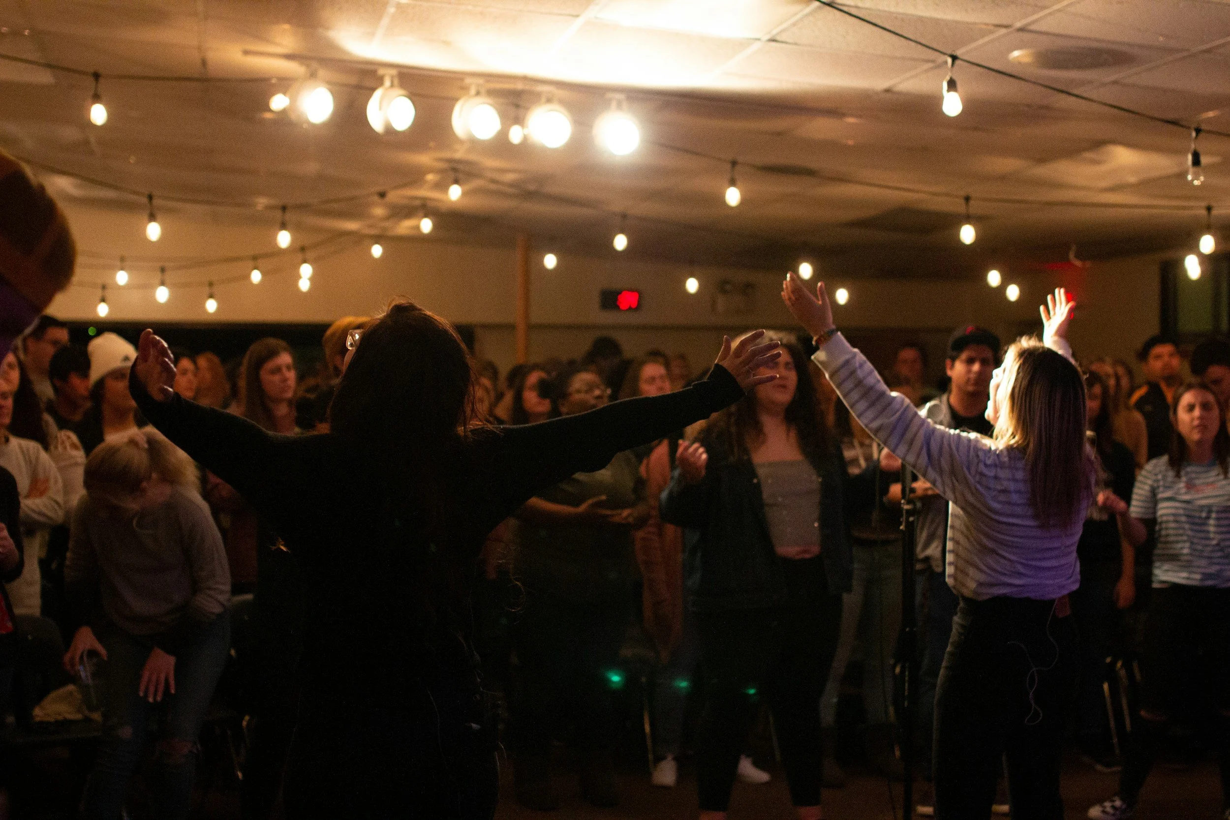 People gathered in a dimly lit indoor space with string lights, participating in a worship or prayer session, with two women at the front raising their arms.