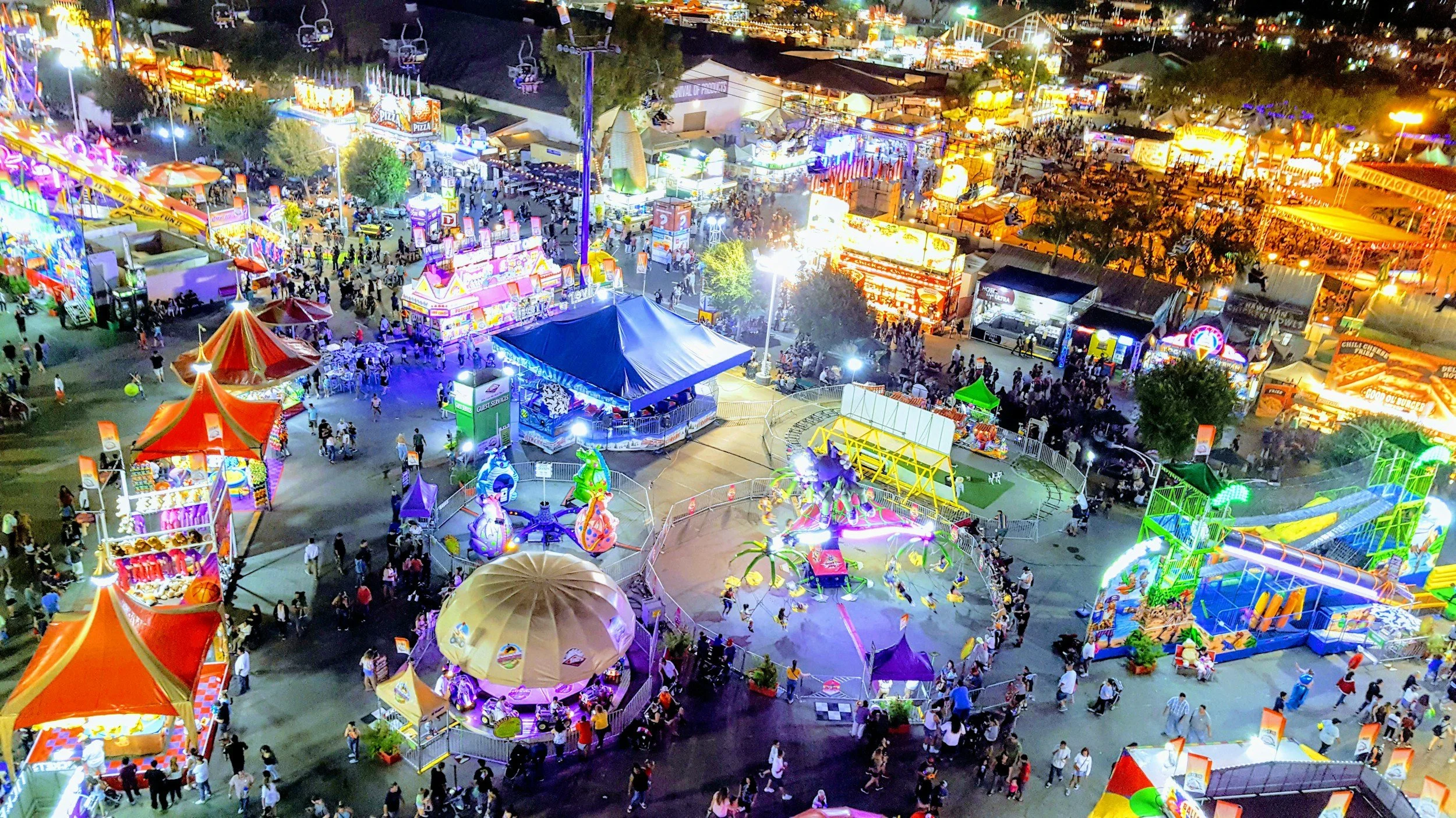 Nighttime view of a bustling carnival with bright lights, rides, games, and crowds of people walking around.