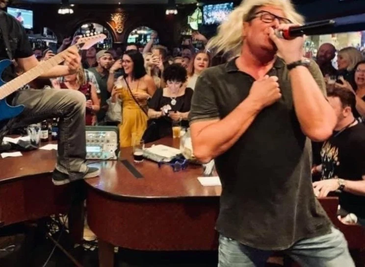 A musician with long blonde hair, wearing glasses and a dark shirt, passionately singing into a microphone in a crowded bar or club with an audience watching and taking photos in las vegas