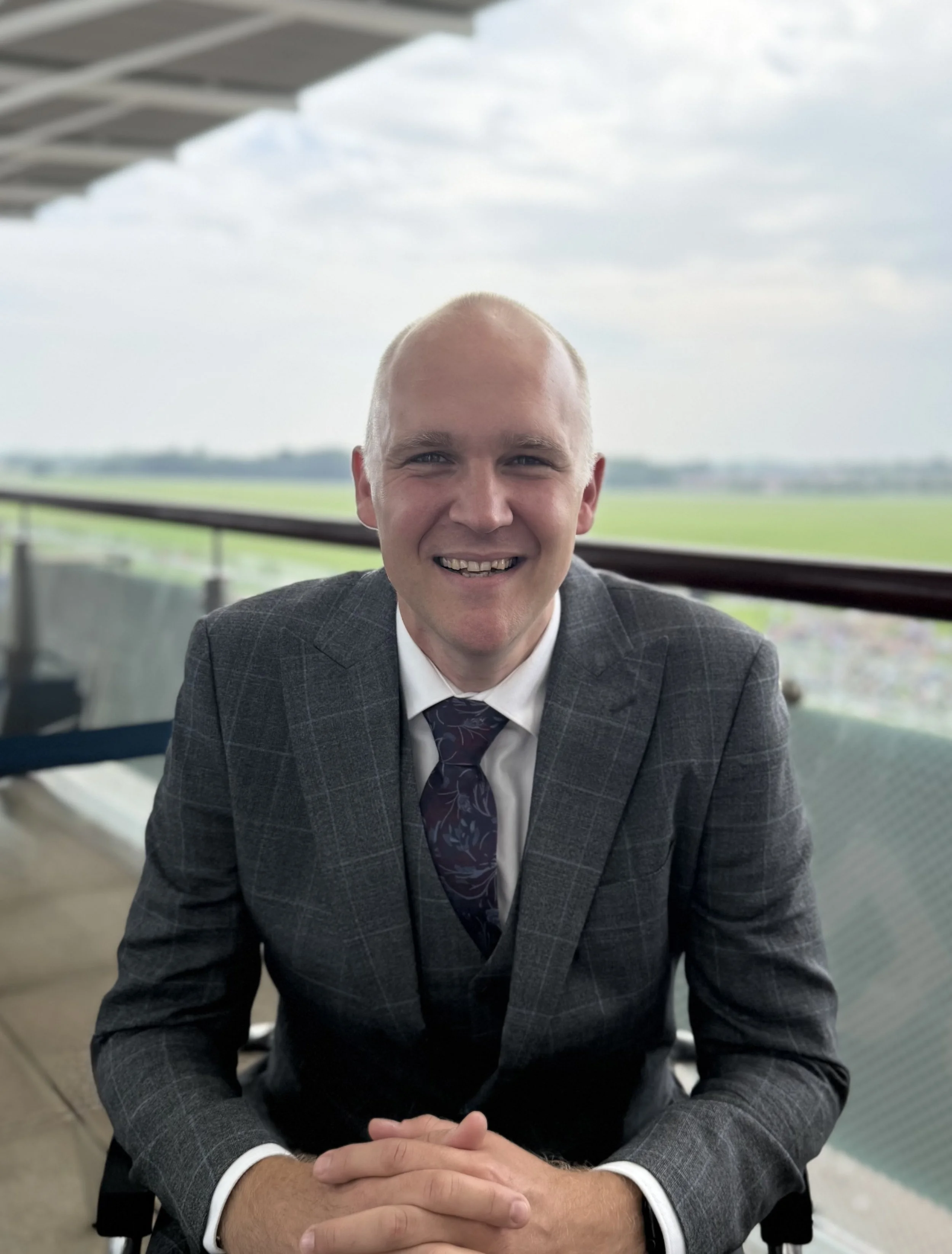 Male with very short grey hair, wearing a grey suit and lilac shirt and purple tie, smiling, looking at the camera