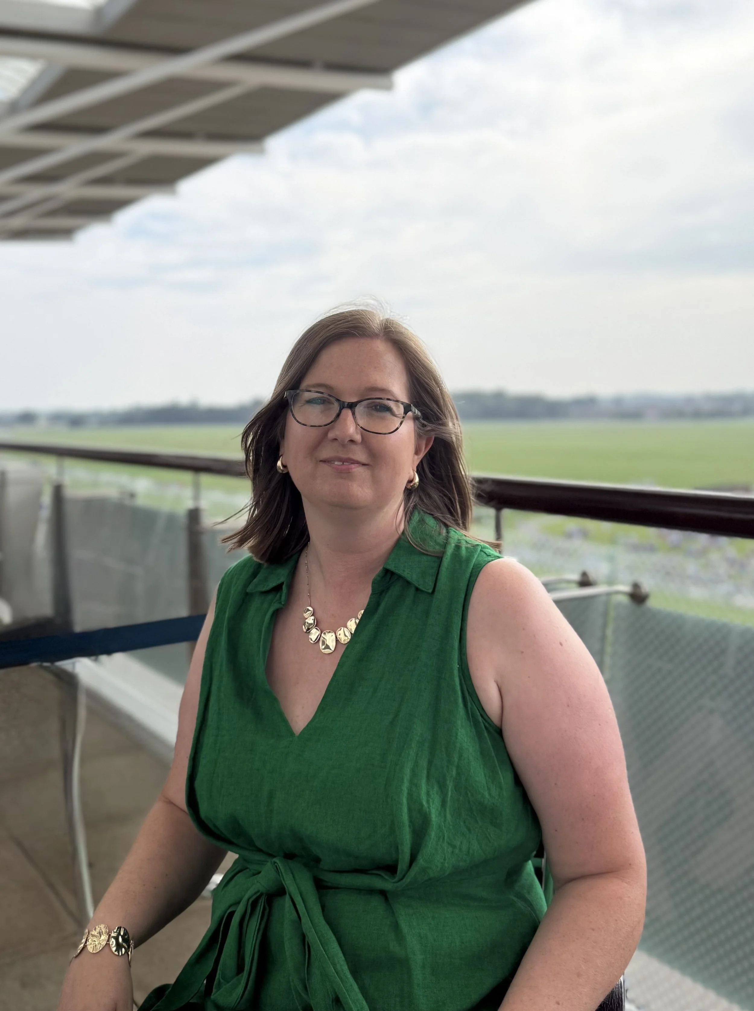 Female with long blonder hair, glasses, wearing a bead necklace and a sleeveless beige top