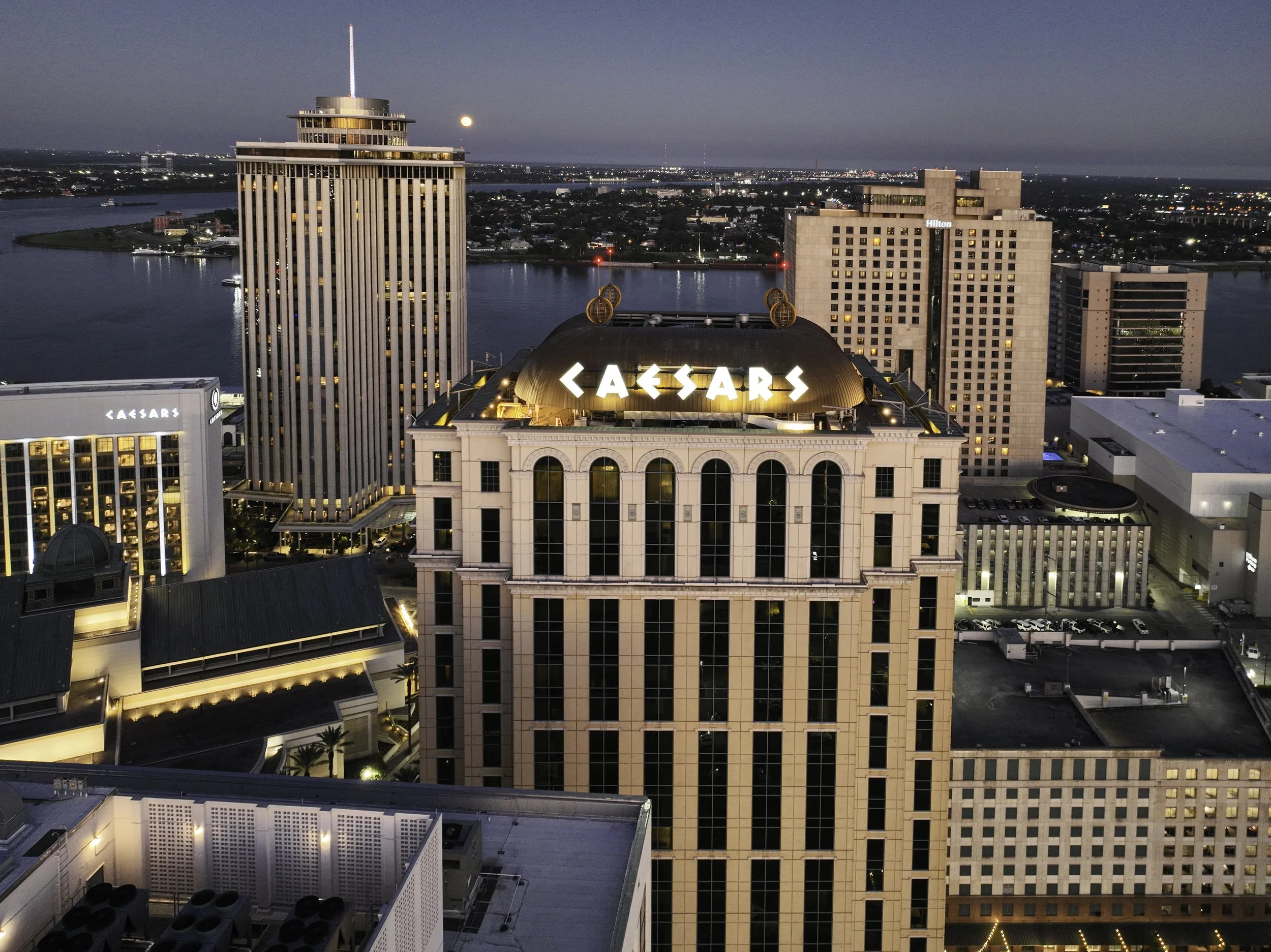 Nighttime aerial view of Caesars Palace hotel and casino in Las Vegas, with the illuminated Caesars sign on top of the building and a river in the background.