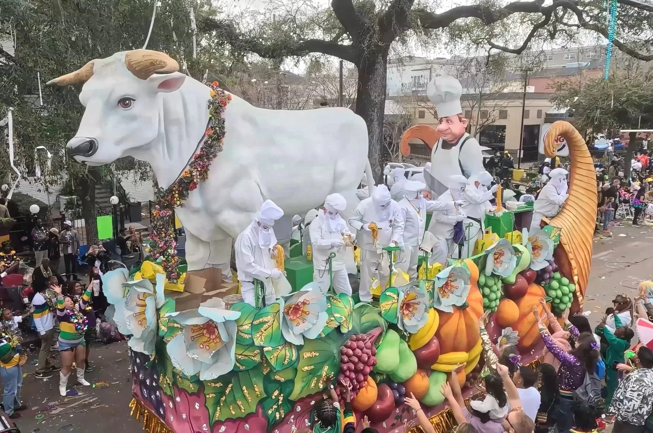 Large parade float decorated with a white cow statue with horns and a floral necklace, and a chef figure, surrounded by people in colorful costumes, with a crowd in the background.