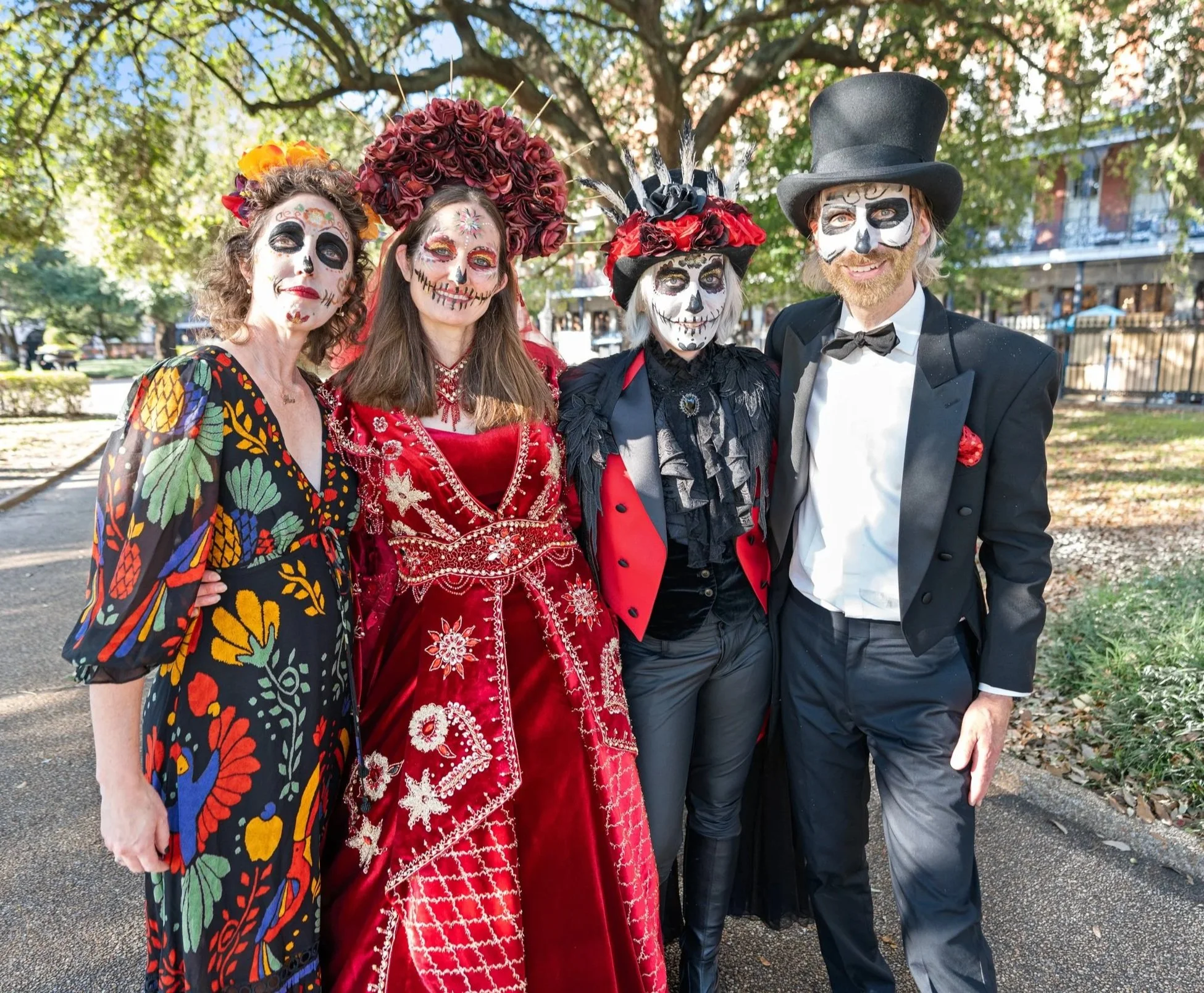 Four people dressed in Día de los Muertos costumes with sugar skull face paint standing outdoors in a park on a sunny day.