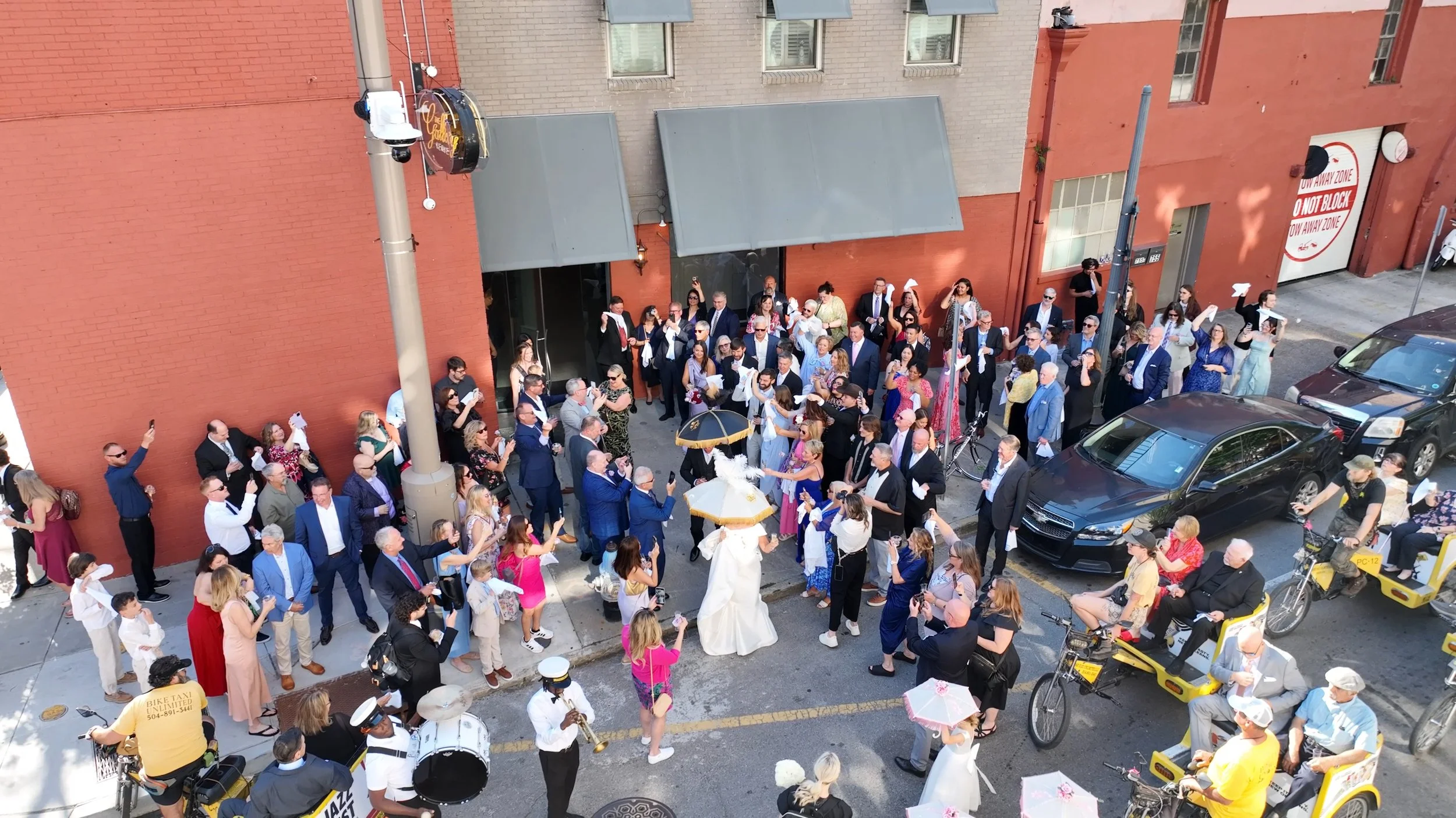 A crowd of people gathered outside a brick building on a city street, some holding umbrellas, with a couple of performers in white costumes in the center, surrounded by police officers, taxis, and cyclists.