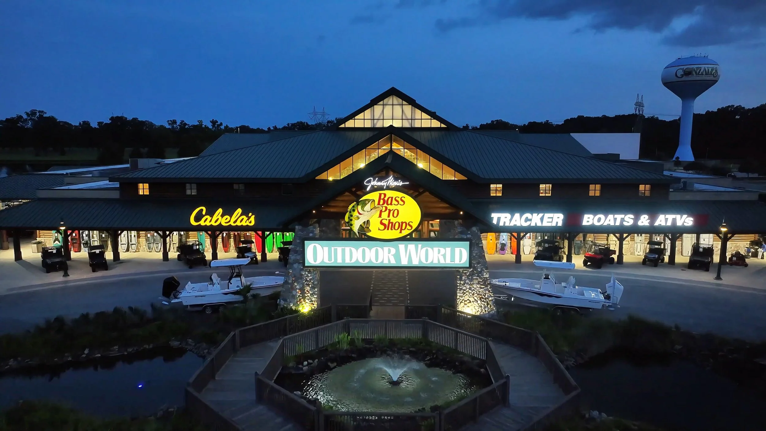 Night view of Outdoor World store with illuminated signs for Cabela's, Bass Pro Shops, Tracker Boats & ATVs, and outdoor gear; boats parked outside and a water fountain in foreground.