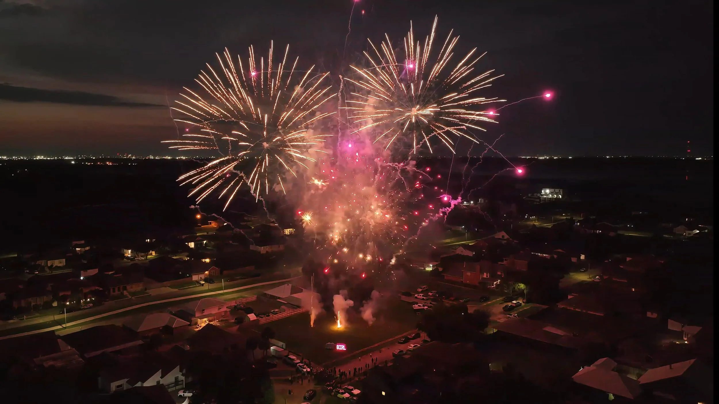 Nighttime fireworks display over a suburban neighborhood with houses, cars, and a crowd gathered below.