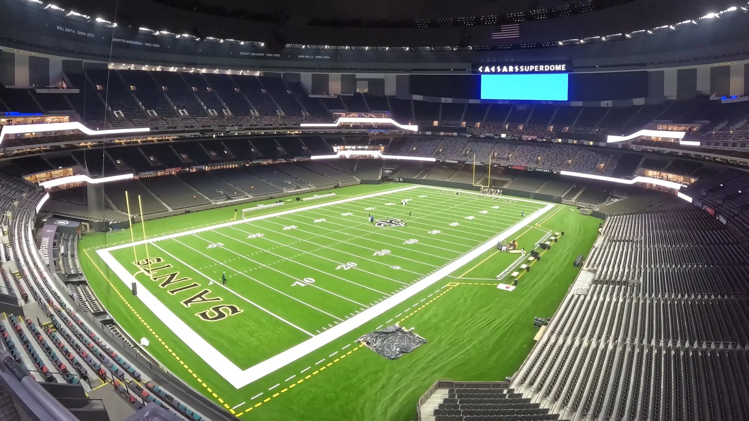 Empty football field inside Caesars Superdome with team logos, seats, and large digital scoreboard.