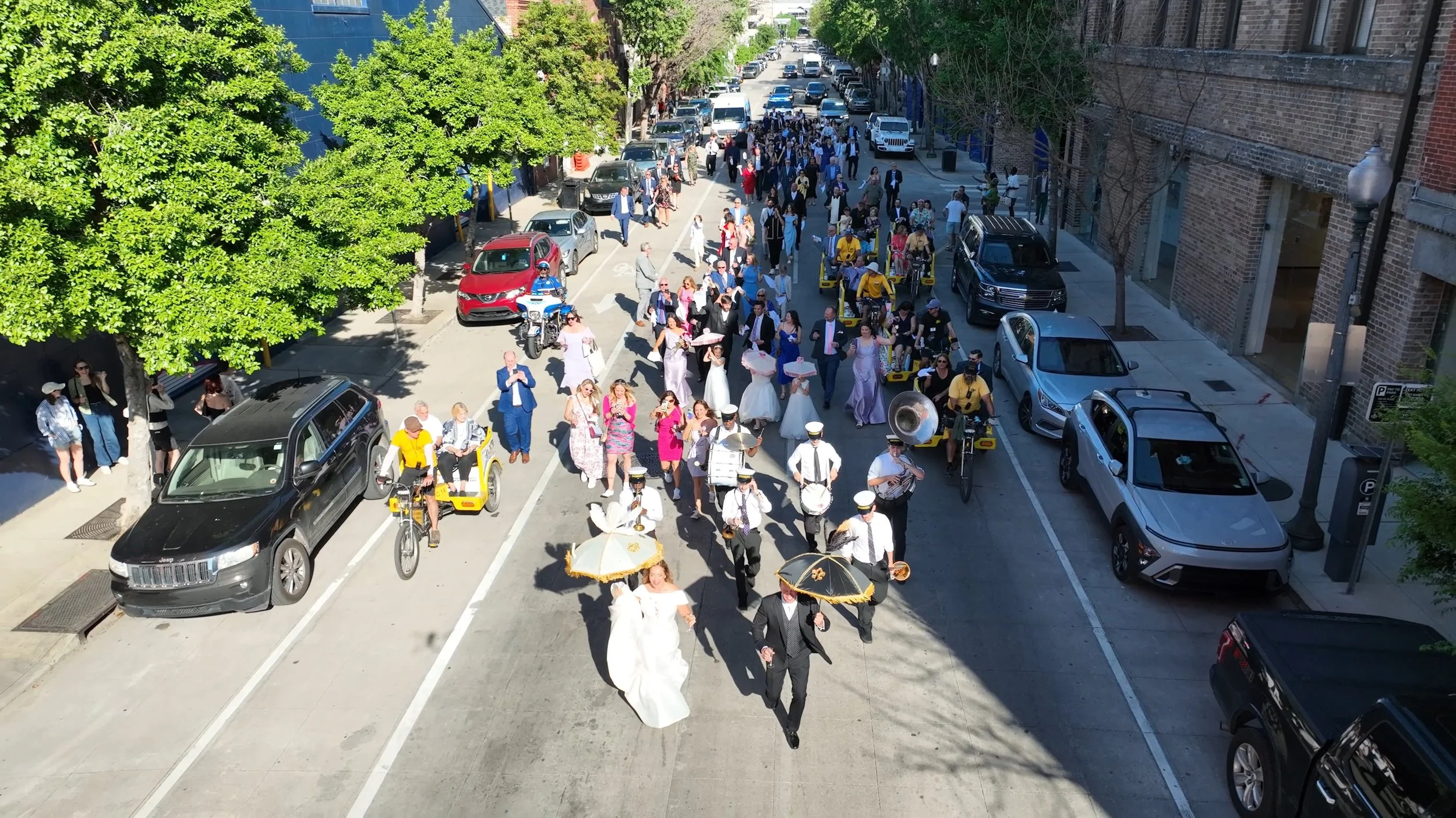 A wedding procession walking down a city street with a bride and groom at the front, holding umbrellas, surrounded by a group of bridesmaids, groomsmen, and guests. The street is lined with parked cars and pedestrians, with trees and buildings along 