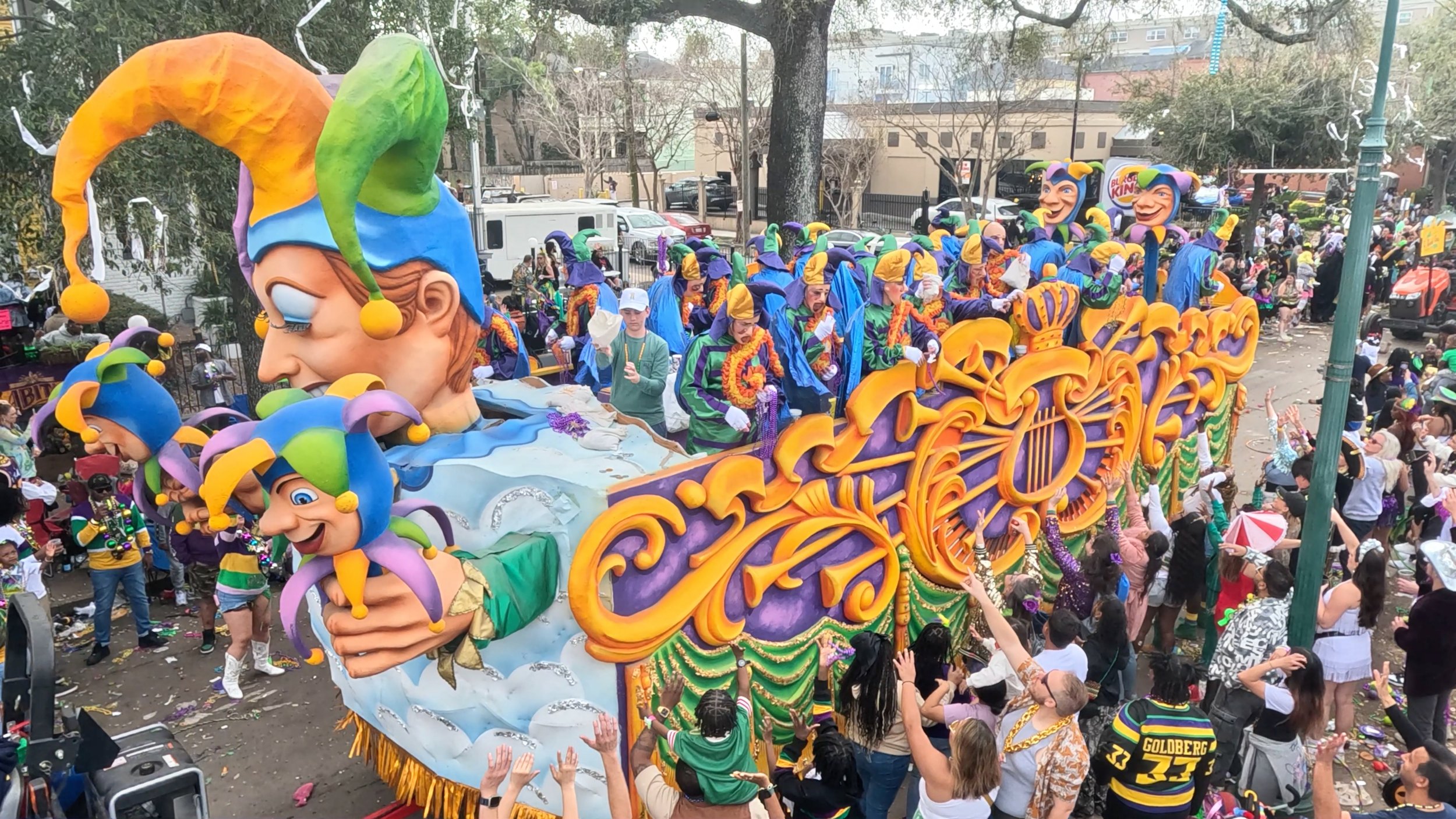 Colorful Mardi Gras parade float with large smiling jester faces, decorated with purple, green, and gold, and a crowd of people celebrating around it in a city street.