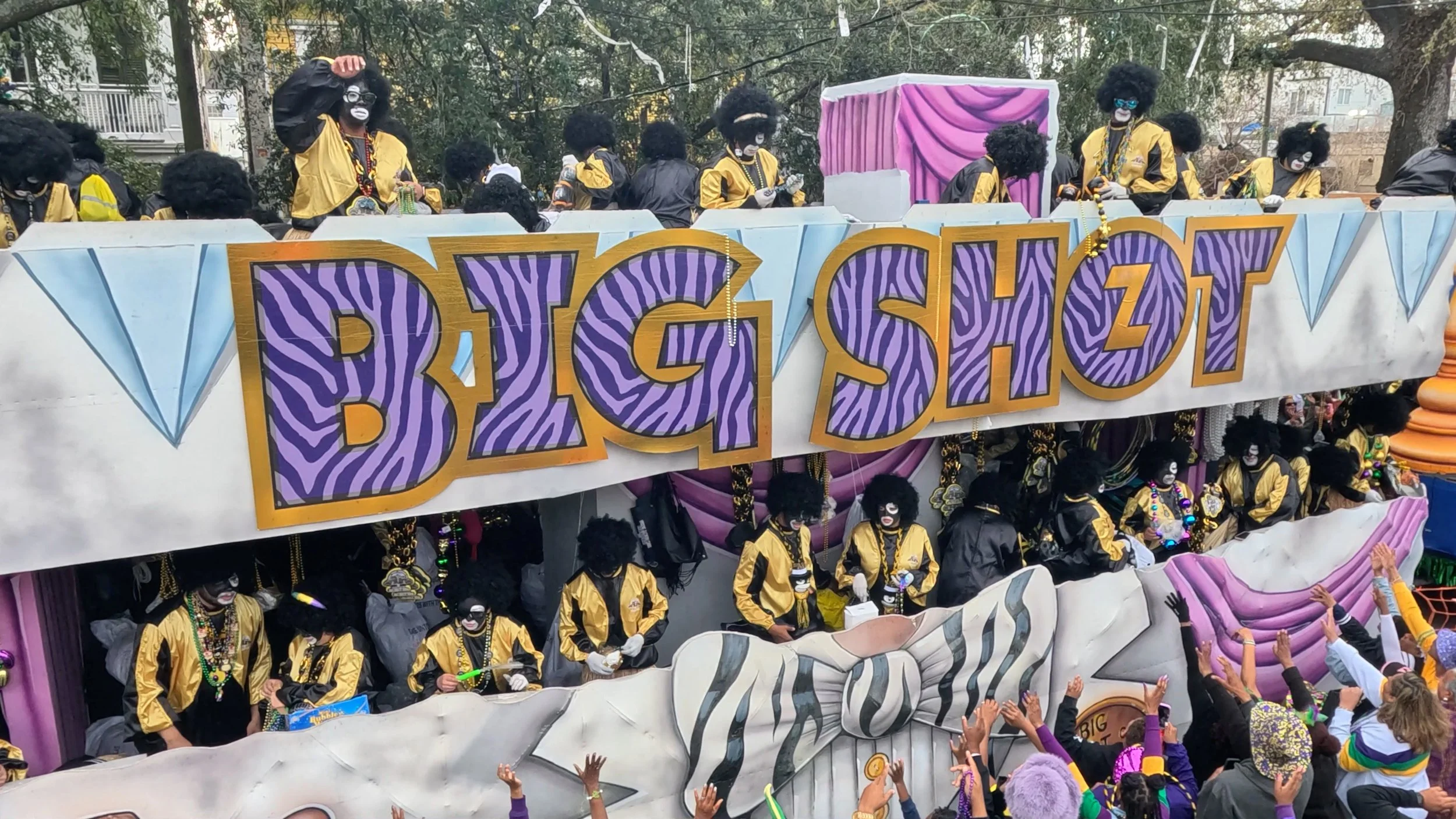A Mardi Gras parade float with people dressed in blackface makeup and costumes, wearing yellow jackets, around a large sign reading 'BIG SHOT' with purple and black zebra stripe pattern and gold lettering, in front of a crowd with hands raised.