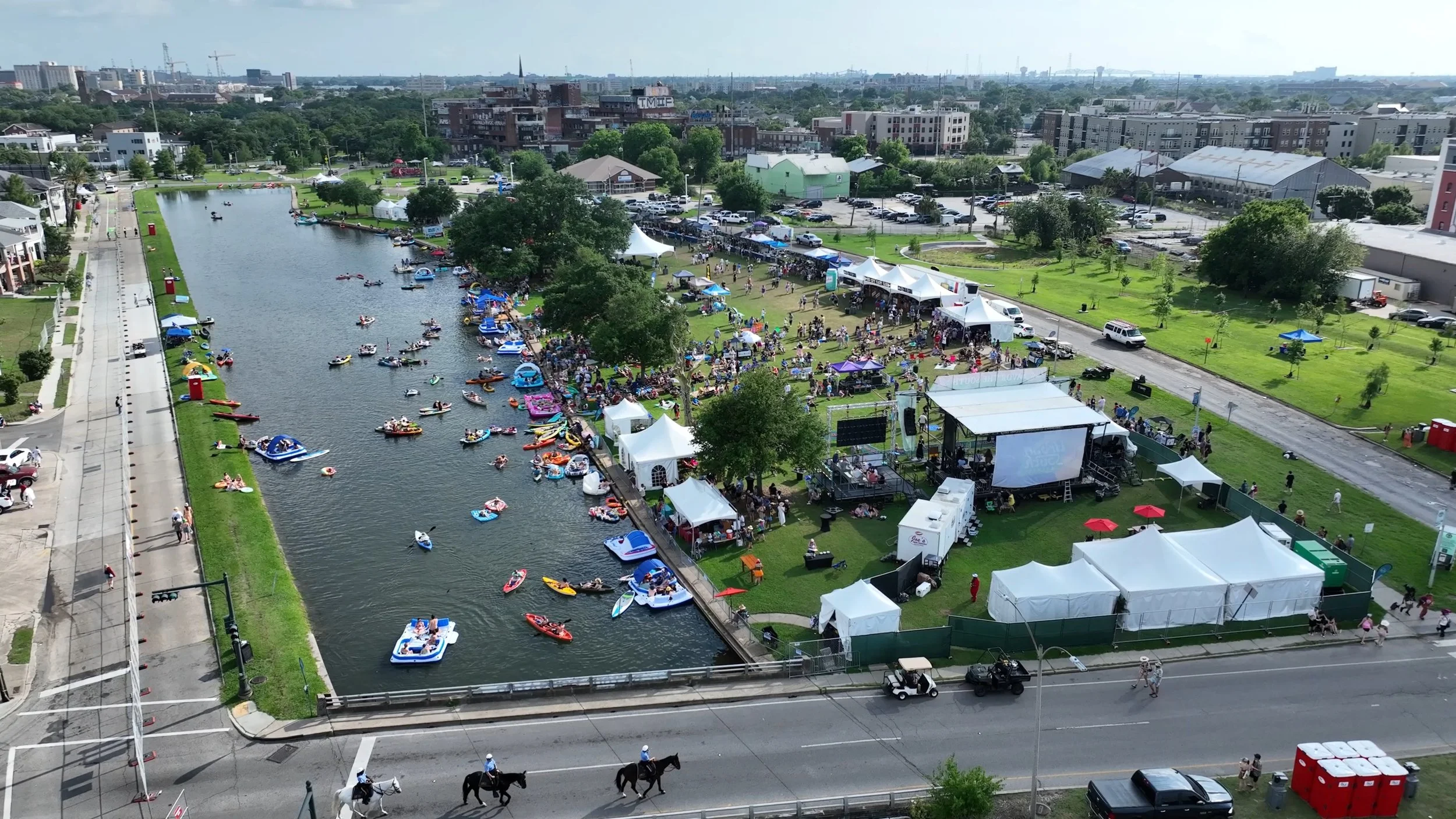 Aerial view of a lively outdoor event by a river with many boats, tents, and a stage, surrounded by city buildings and green space.