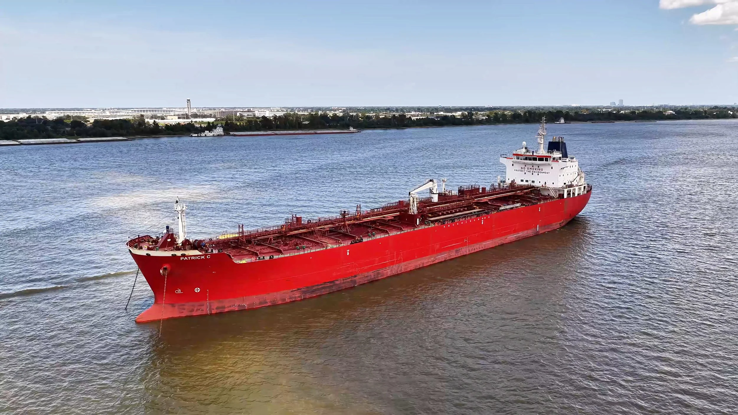 A red cargo ship named Patrick C sailing on water with a distant shoreline and cityscape in the background.