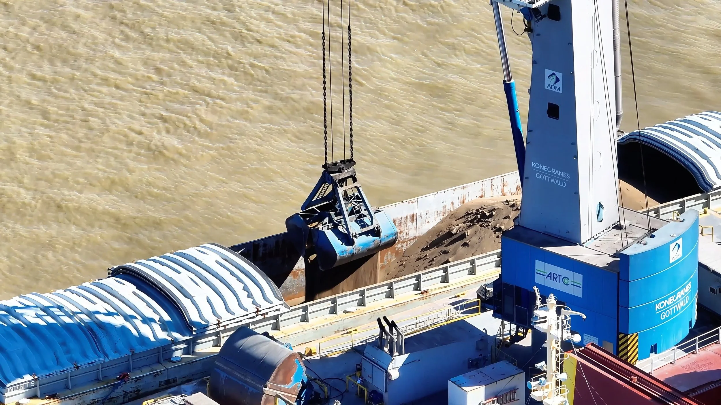 A large industrial dredging vessel with a blue crane lifting a bucket of sediment from a river or harbor, with muddy water in the background.