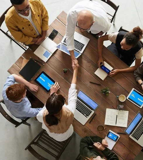 A group of six people engaged in a meeting around a wooden table with laptops, tablets, notebooks, and a cup of coffee, shaking hands.
