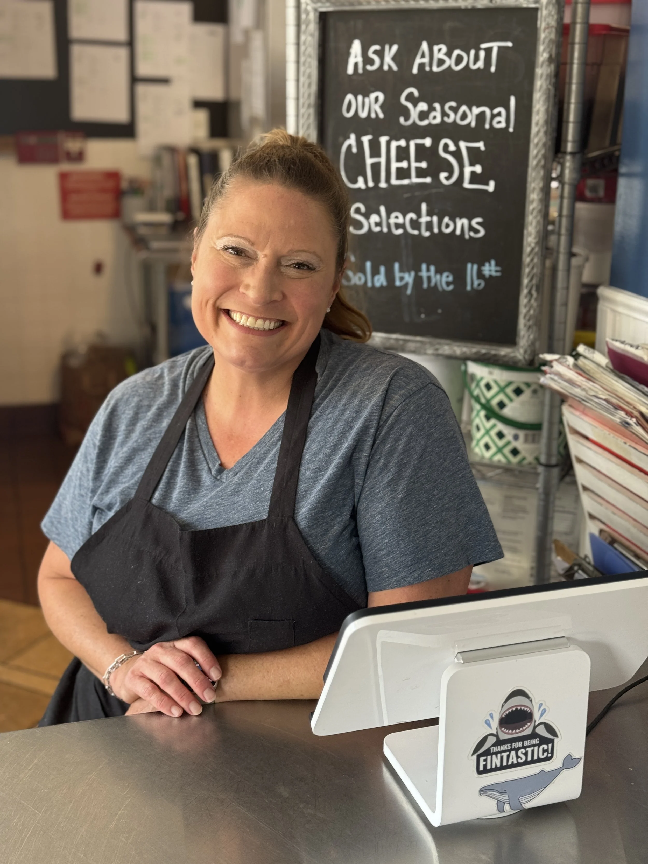 woman smiling in a black apron behind the register in a shop with a cheese sign behind her in a a blue shirt with a ponytail