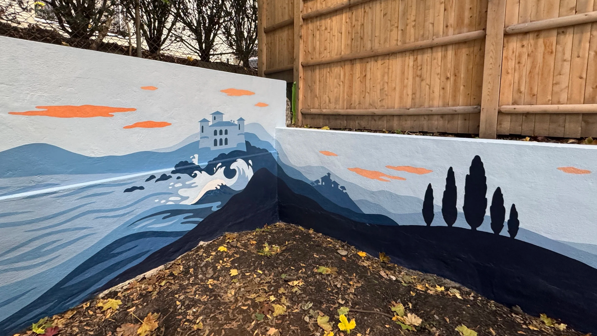 A blue monochrome graphic wallpaper-inspired mural showing a lake with waves crashing against a landscape with rolling hills, buildings, and Italian plants, painted across two walls that meet in a corner in a backyard in Boston